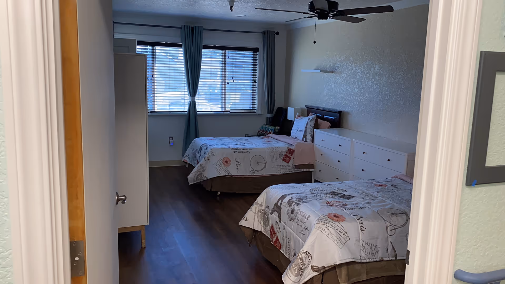 A bedroom with two twin beds covered in Paris-themed bedspreads. The room has a wooden floor, a white dresser between the beds, a window with blue curtains and blinds, and a ceiling fan above.
