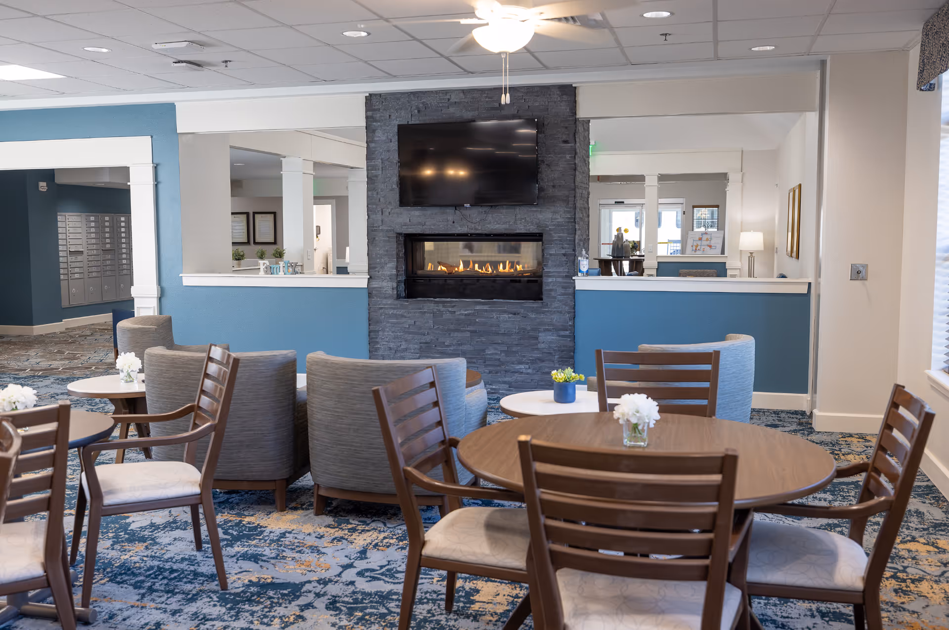 A cozy common area in a senior living facility featuring a modern gas fireplace set in a dark stone wall with a flat-screen TV mounted above it. The room has several round wooden tables with chairs and small flower arrangements on top. Comfortable gray armchairs are arranged around the fireplace. The walls are painted blue and white, and the carpet has a blue and beige pattern. There is a ceiling fan above and a window with blinds on the right side.