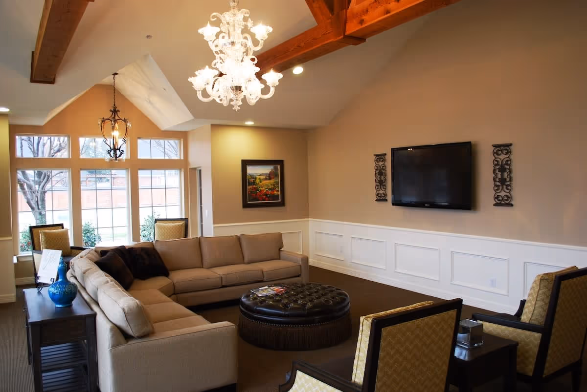 A bright communal living room with a beige sectional sofa, round tufted ottoman, wall-mounted TV, and large windows.