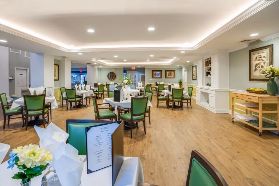 A spacious and well-lit dining room with multiple tables covered with white tablecloths and set with napkins and menus. The chairs have green cushions, and there are decorative flowers on the tables. The room has wooden flooring, light-colored walls with framed artwork, and recessed ceiling lights. A wooden side table with a green vase and flowers is visible on the right side.