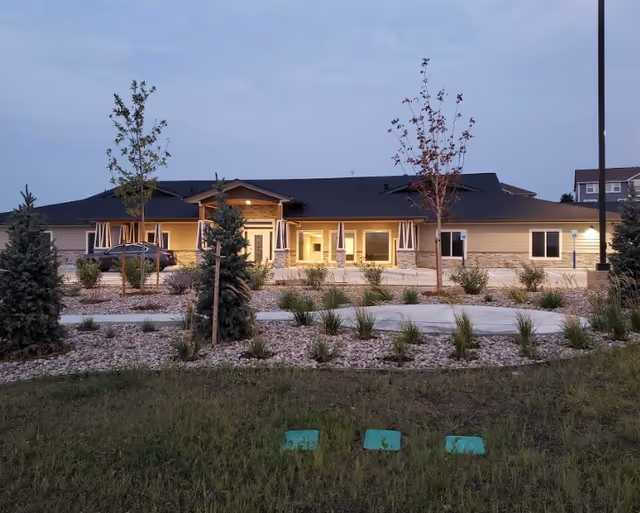 Exterior view of a single-story assisted living facility building at dusk with illuminated windows and entrance. The building is surrounded by landscaped grounds with small trees, shrubs, and a paved walkway.