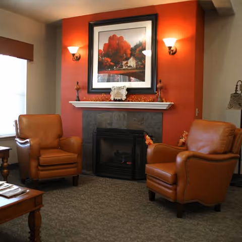 A cozy living room area with two brown leather armchairs facing each other near a fireplace with a dark tile surround. Above the fireplace is a white mantel decorated with small items and a framed picture of a house with red trees. The wall behind the fireplace is painted red, and there are two wall sconces providing warm light. A window with blinds is visible on the left side.