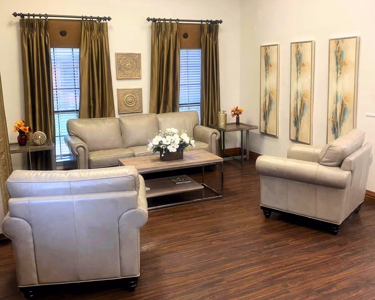 A cozy living room with a beige leather sofa and two matching armchairs arranged around a wooden coffee table with a floral centerpiece. The room features dark wood flooring, two windows with brown curtains, decorative wall art, and side tables with vases of flowers.