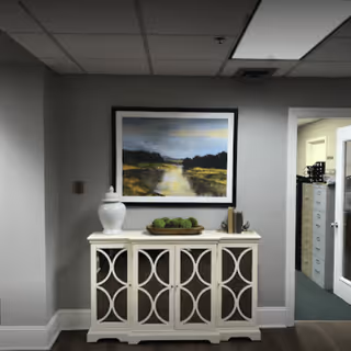 A hallway area in a healthcare facility with a white decorative cabinet featuring circular patterns on the doors. On top of the cabinet are a white vase, a tray with green decorative spheres, and some books. Above the cabinet hangs a framed landscape painting depicting a river or marsh with trees and a cloudy sky. To the right, there is an open doorway leading to an office area with file cabinets and a door with glass panels.