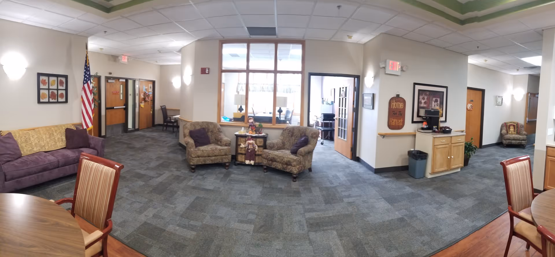 A spacious senior living facility common area with multiple seating options including a purple couch, two patterned armchairs, and a single armchair in the hallway. There is an American flag near a set of double doors, framed artwork on the walls, and a small table with decorative items between the armchairs. The room has carpeted flooring, a drop ceiling with fluorescent lighting, and a small wooden cabinet with a computer station. The area appears clean and welcoming with neutral wall colors and natural light coming through an interior window.