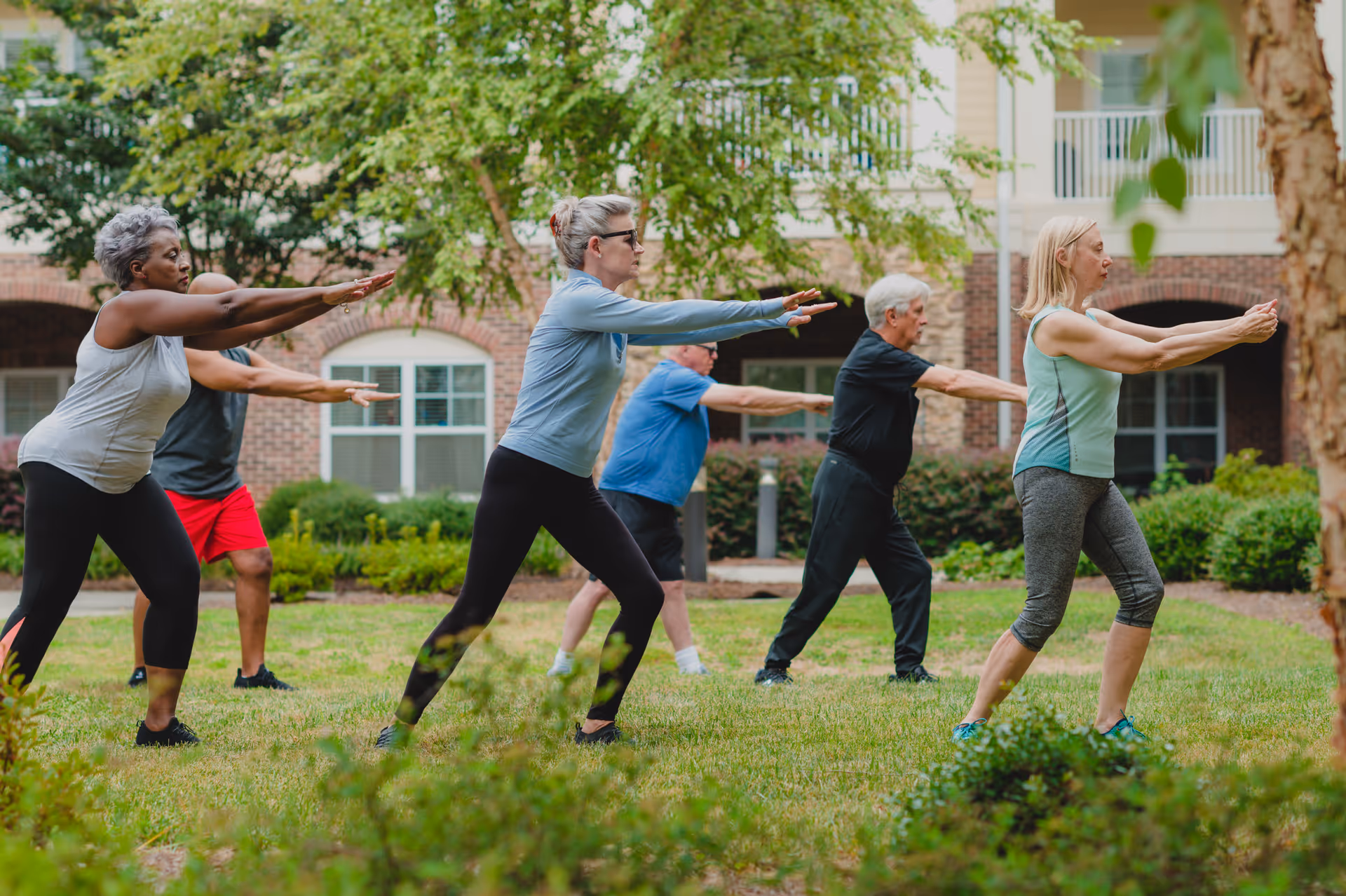 A group of older adults participating in an outdoor exercise class on a grassy area in front of a brick building with windows and greenery. They are all standing with arms extended forward, engaged in a stretching or balance exercise.