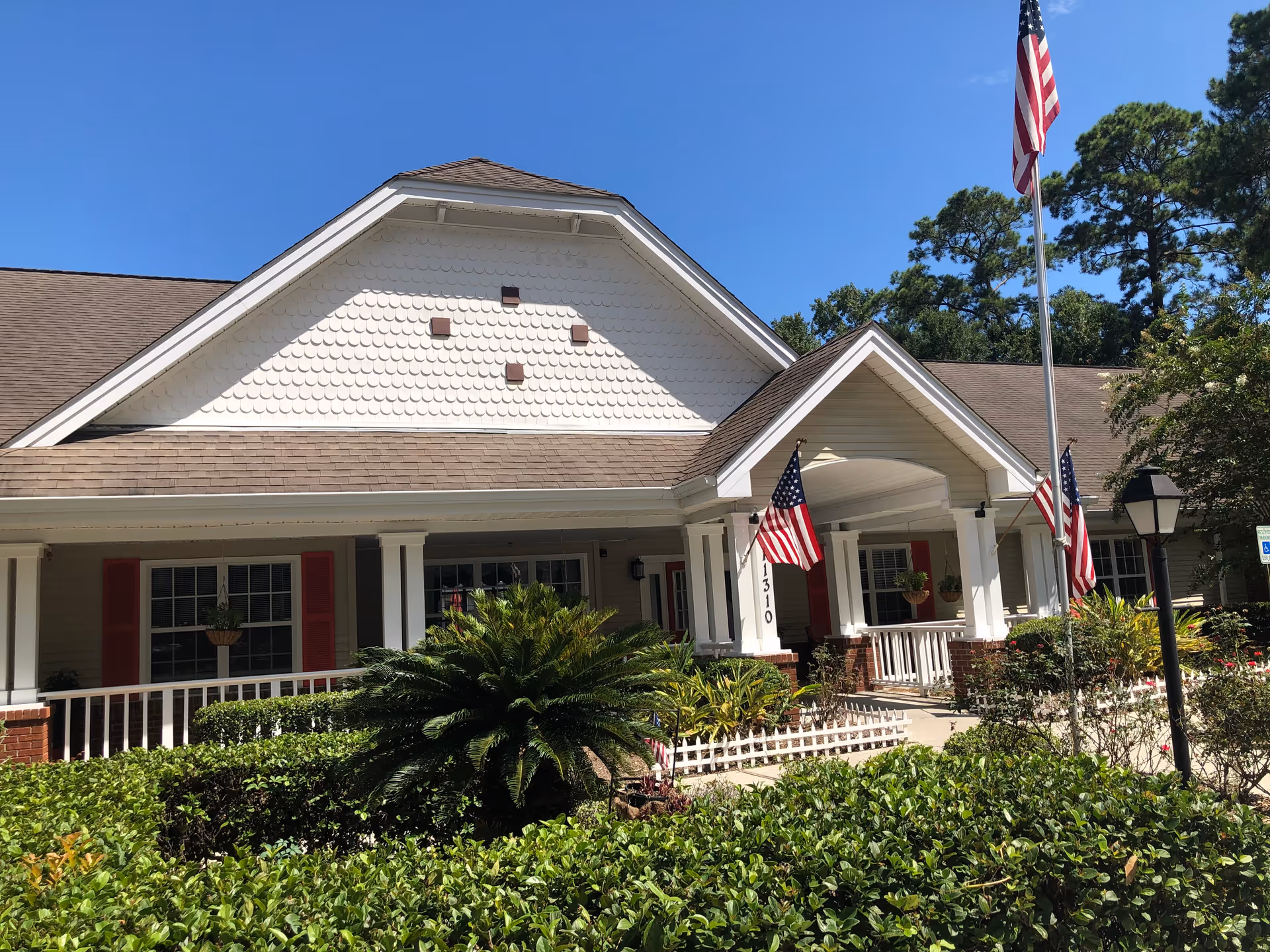Front exterior view of a single-story building with a covered porch, white columns, and red shutters. The building is surrounded by green shrubs and plants, with three American flags displayed near the entrance and a flagpole with an American flag in front. The sky is clear and blue.