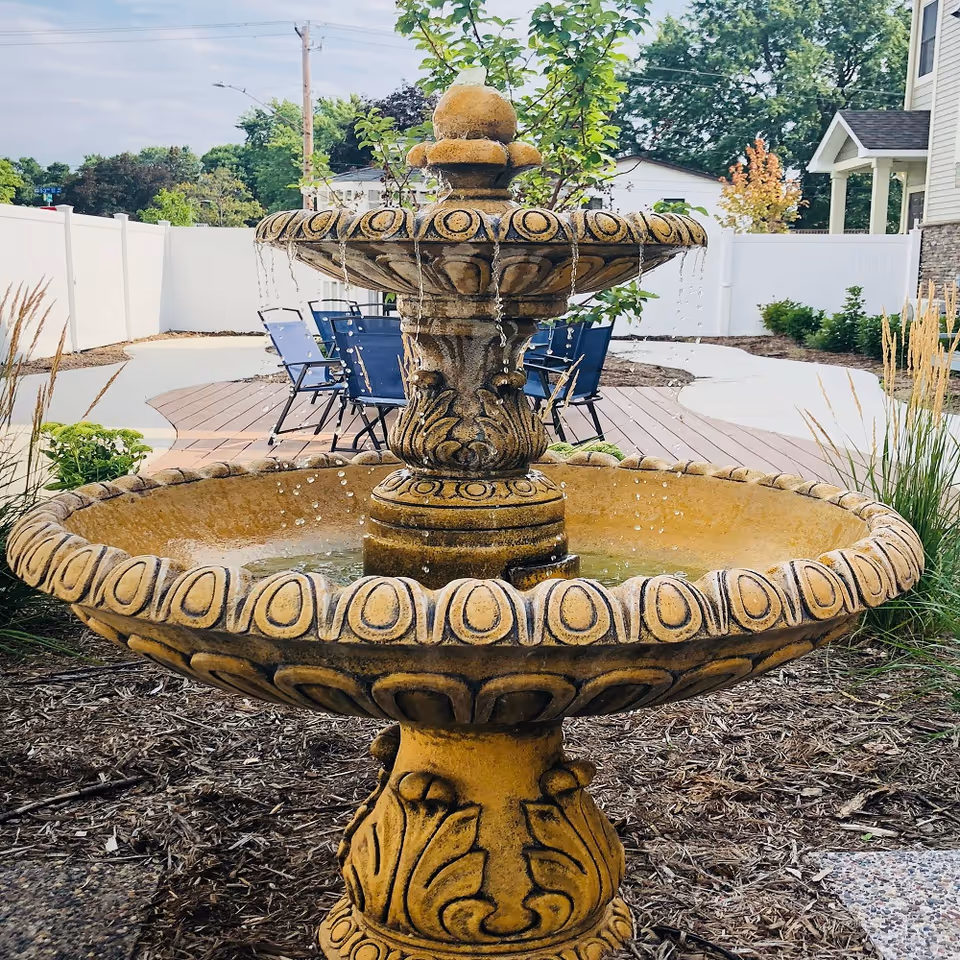 A decorative two-tiered stone water fountain with water streams flowing from the upper tier to the lower basin, set in an outdoor garden area with mulch, plants, a white fence, and blue patio chairs in the background.