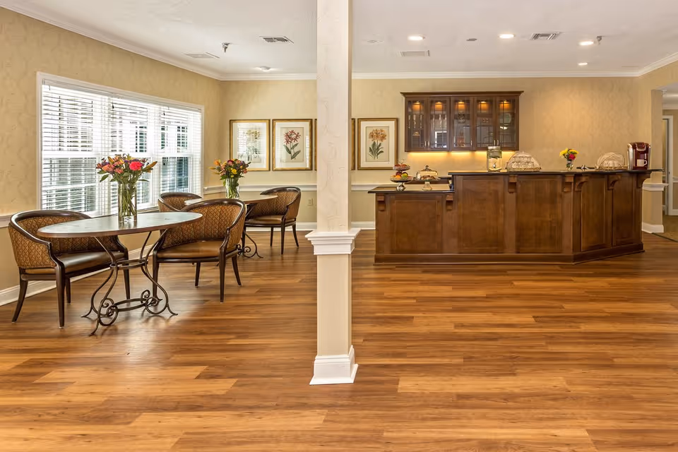 Bright communal dining area with wooden floors, round tables and chairs, flower centerpieces, and a wooden service counter.