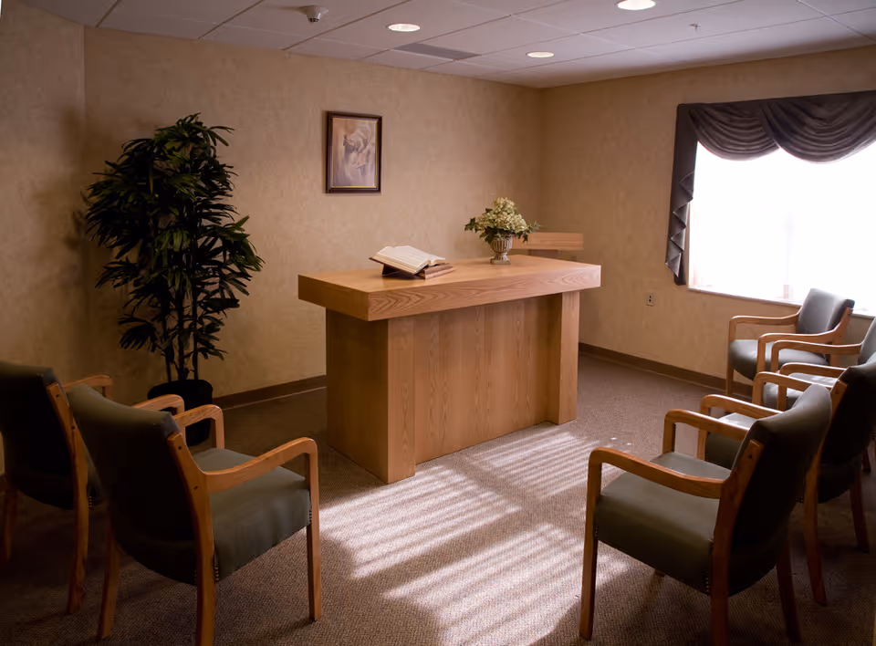 A small room with six wooden chairs arranged in two rows facing a wooden podium with an open book and a flower vase on top. There is a framed picture on the wall and a large window with drapes letting in sunlight. A tall potted plant is placed in the corner.