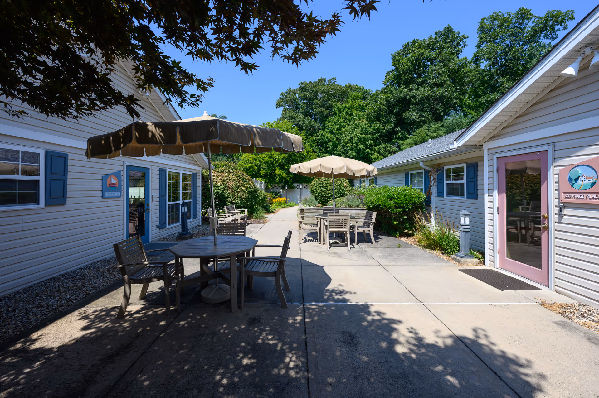 Outdoor patio area between two single-story buildings with beige siding and blue shutters. The patio has round tables with umbrellas and wooden chairs. There are bushes and trees surrounding the area under a clear blue sky.