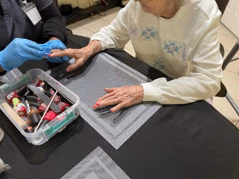 An elderly woman wearing a white sweater is having her nails painted by a person wearing blue gloves. Various nail polish bottles and manicure tools are on the table covered with a black cloth and gray placemats.