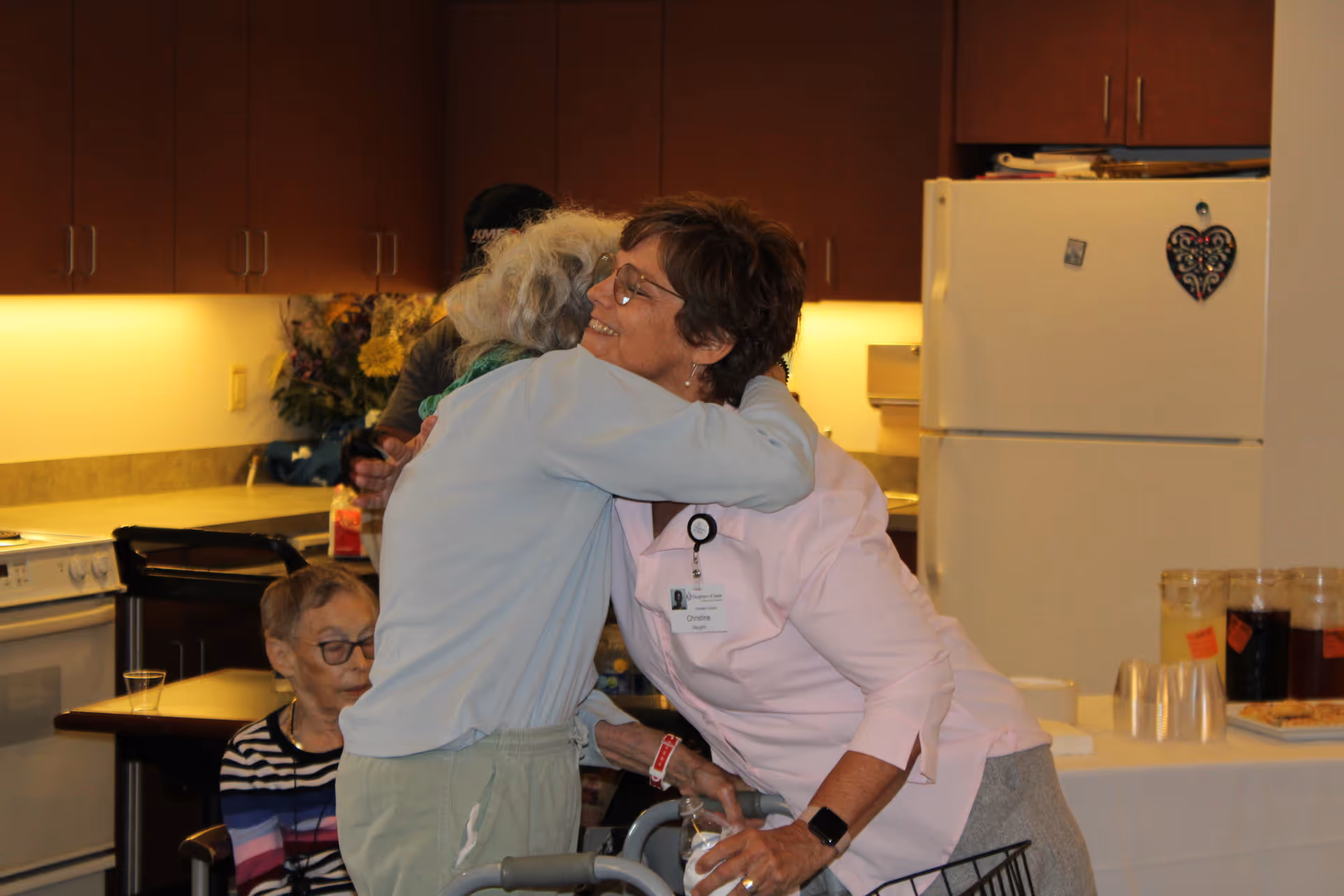 Two women hugging in a kitchen area of an assisted living facility. One woman is wearing a light blue top and using a walker, while the other woman, wearing glasses and a pink shirt with a name badge, smiles warmly. In the background, another elderly woman is seated at a table. The kitchen has wooden cabinets, a white refrigerator with magnets, and a counter with pitchers and cups.