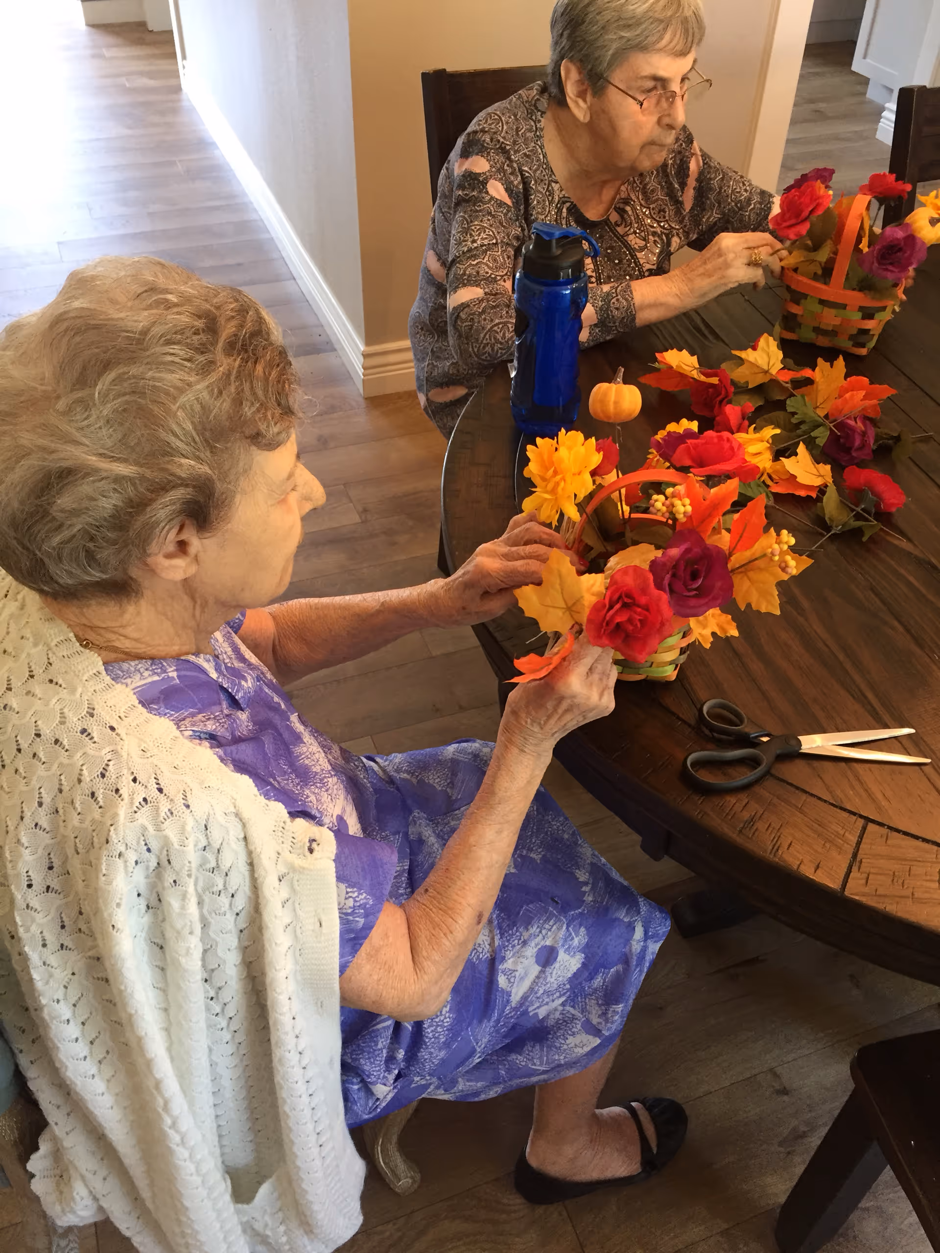Two elderly women sitting at a wooden table engaged in a craft activity, arranging colorful artificial flowers and autumn leaves in small woven baskets. One woman is wearing a purple dress with a white knitted shawl, and the other is wearing glasses and a patterned top. A pair of scissors and a blue water bottle are also on the table.
