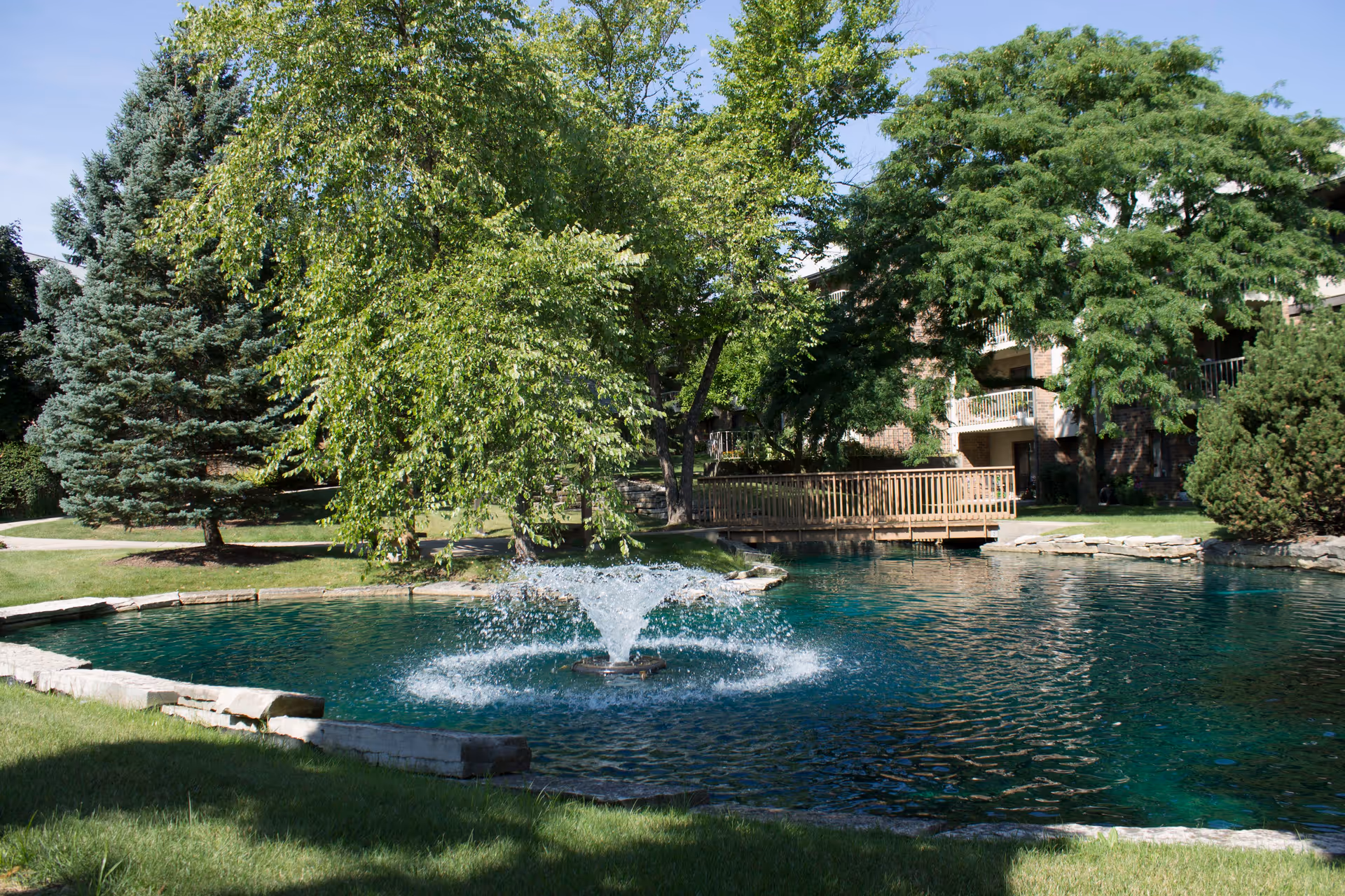 A serene outdoor scene at VMP Healthcare and Community Living featuring a small pond with a water fountain in the center, surrounded by green grass, trees, and a wooden bridge. In the background, there is a multi-story building partially visible behind the trees.