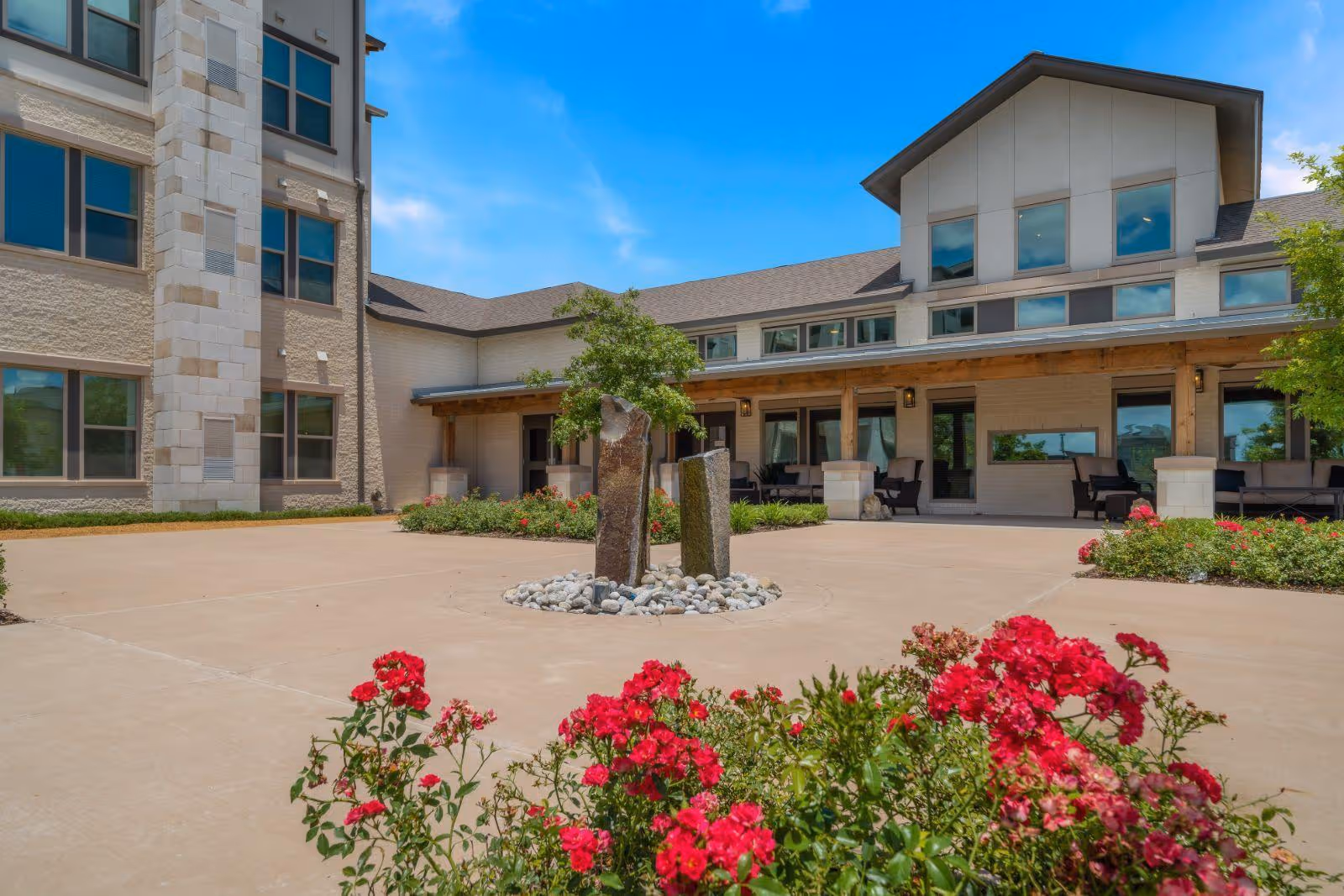 Courtyard and front facade of a senior living building with a small stone fountain, covered seating area, and red flowers in the foreground.