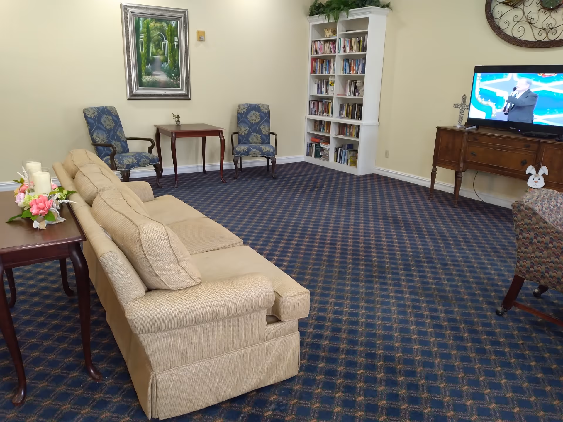 A cozy living room with a beige sofa, two blue patterned armchairs, a wooden side table with candles and flowers, a small wooden table between the armchairs, a white bookshelf filled with books, and a wooden TV stand with a television showing a man speaking. The room has a blue patterned carpet and light yellow walls with a framed painting and a decorative wall clock.