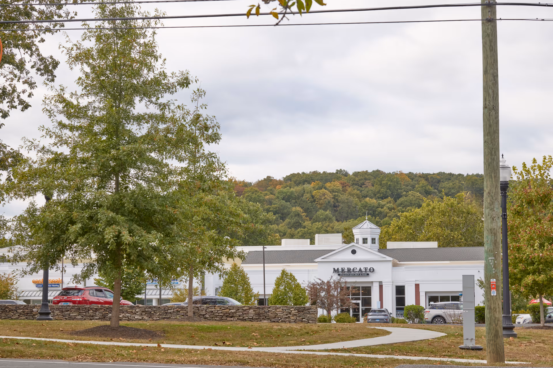 Exterior view of a commercial building named Mercato with a parking lot and several cars in front. There are trees and a stone wall in the foreground, with a wooded hill in the background under a cloudy sky.