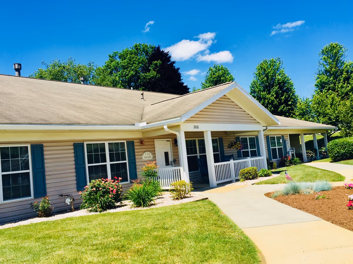 Exterior view of a single-story senior living community building with beige siding, blue shutters, and a covered porch. The building is surrounded by well-maintained landscaping including green grass, bushes, flowers, and trees under a clear blue sky.