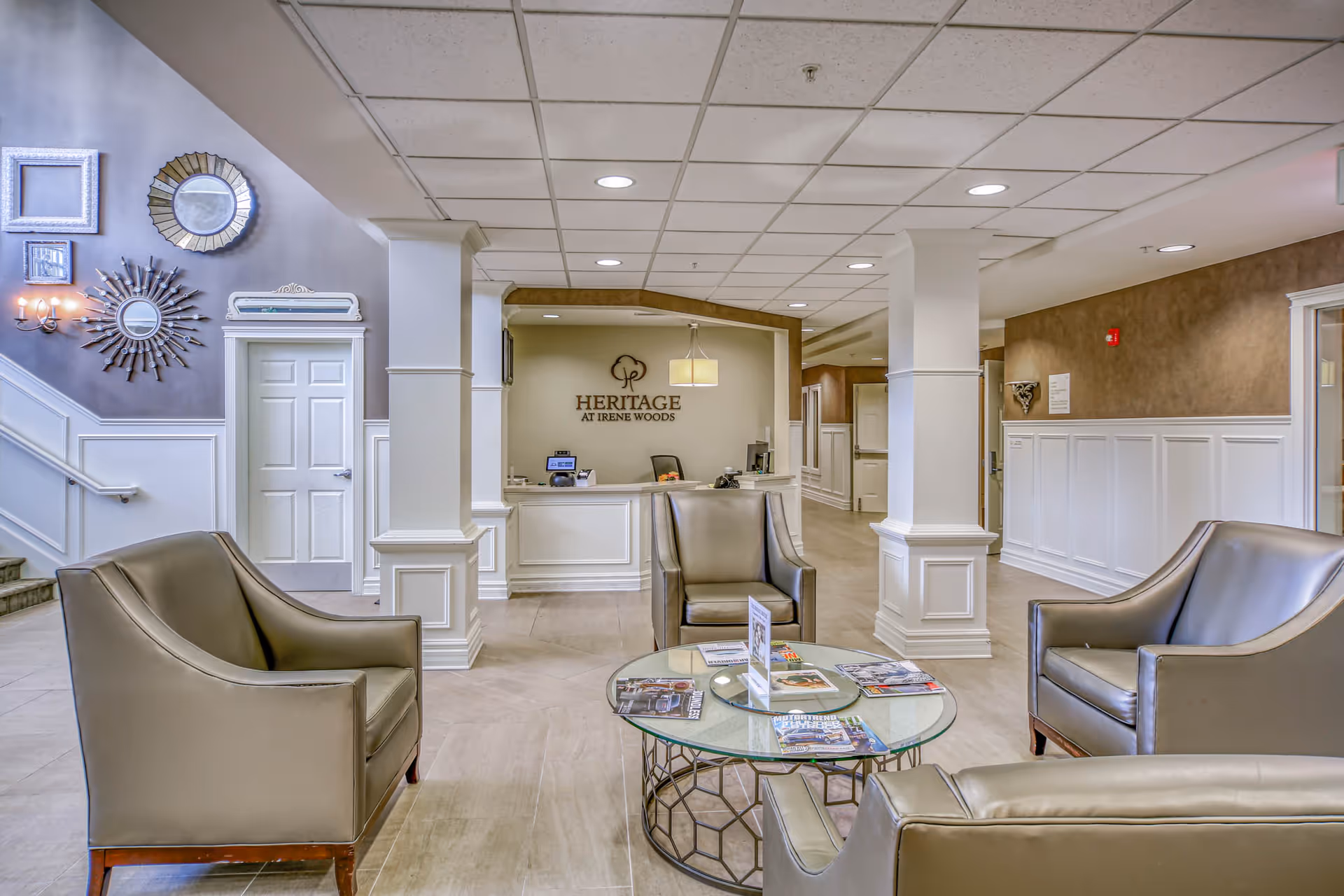 Bright assisted living lobby with leather chairs around a glass coffee table and a reception desk reading 'Heritage at Irene Woods'.