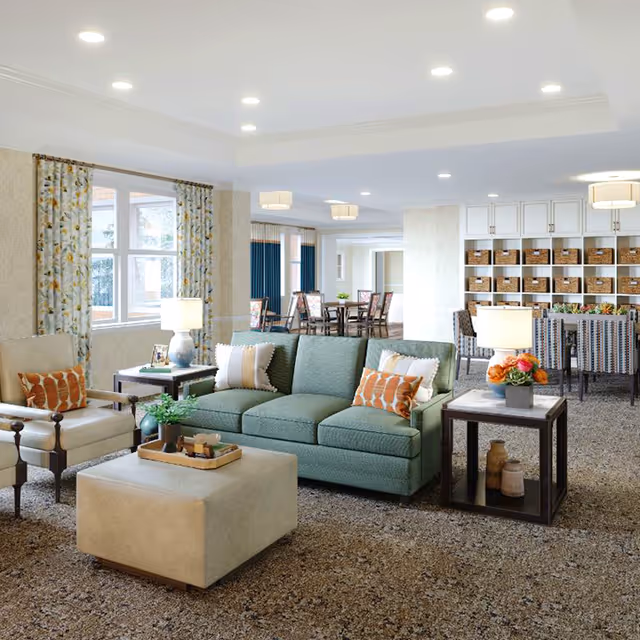 A bright and spacious living room area in a senior living facility featuring a green sofa with decorative pillows, a beige armchair, a square beige ottoman, and two side tables with lamps and floral arrangements. In the background, there is a dining area with tables and chairs, and a shelving unit with baskets. Large windows with patterned curtains allow natural light to fill the room.