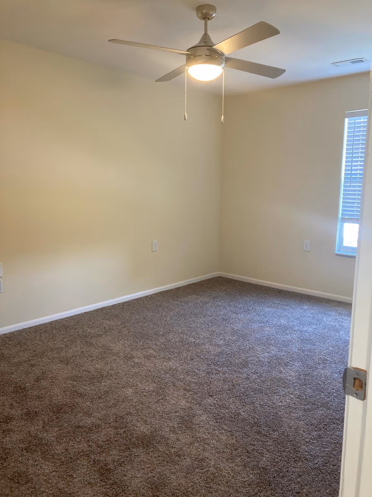 Empty room with beige walls, brown carpet, a ceiling fan with light, and a window with blinds partially open.
