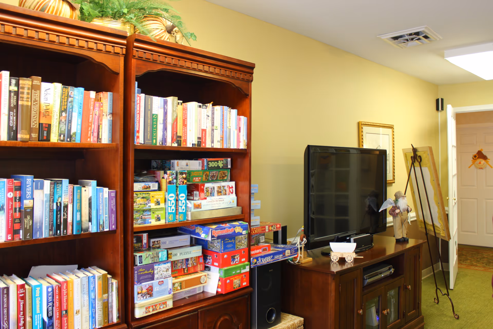 A cozy room in a senior living facility featuring two wooden bookshelves filled with books and board games. To the right, there is a wooden TV stand with a flat-screen television, decorative items, and a framed picture on the wall. The room has light yellow walls and a green carpet, with an open door leading to another area.