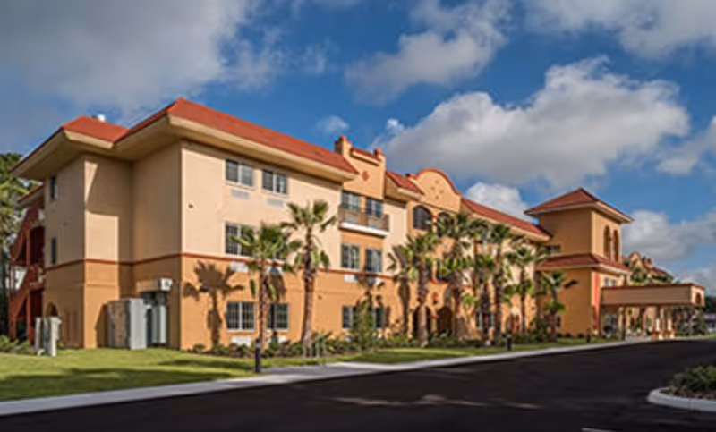Front exterior of a three-story Mediterranean-style senior living building with palm trees and a driveway under a blue sky.