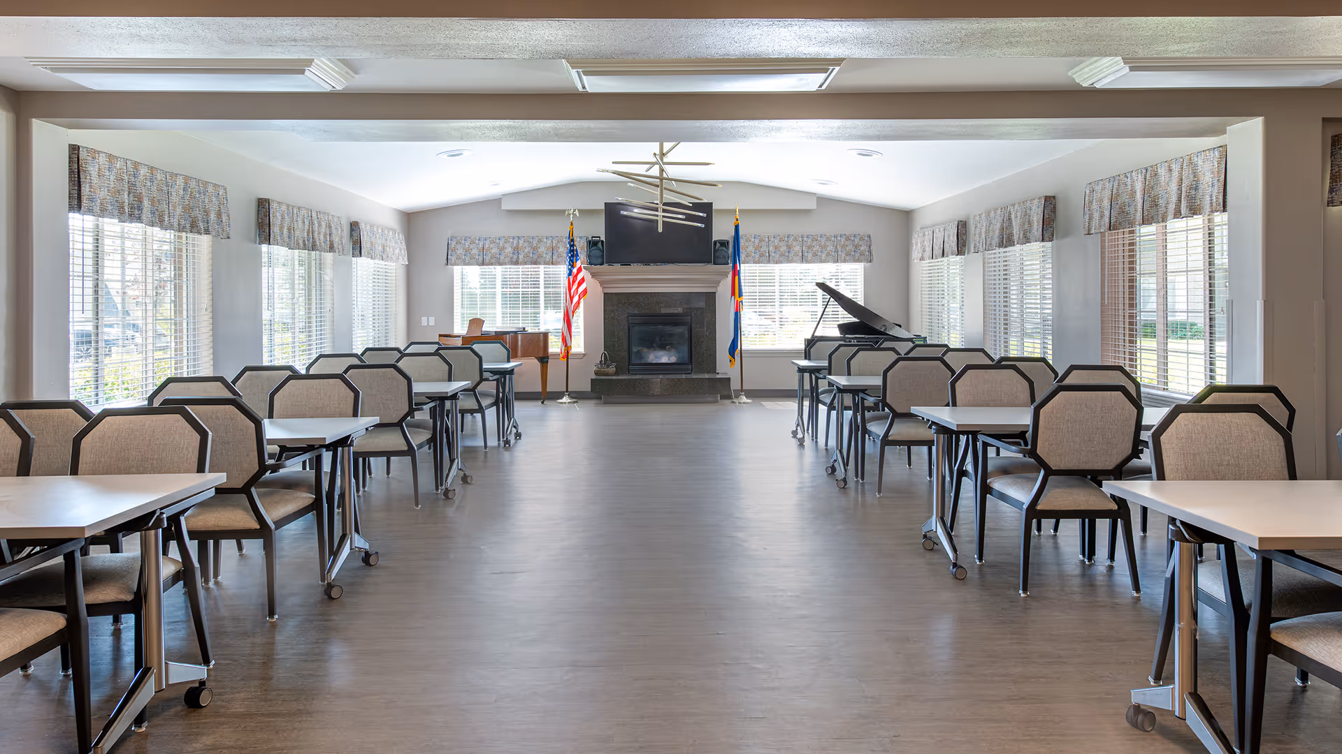 A spacious, well-lit room with multiple tables and chairs arranged in rows along both sides. Large windows with patterned valances allow natural light to fill the room. At the far end, there is a fireplace with an American flag and another flag on either side, a mounted TV above the fireplace, and a grand piano to the right.