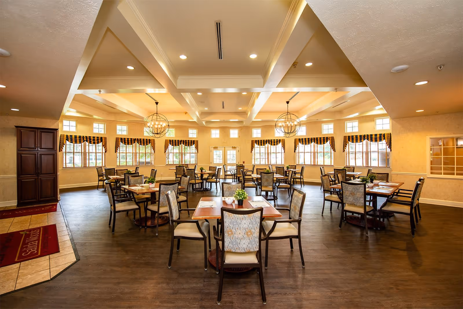 A spacious dining room with multiple wooden tables and cushioned chairs arranged neatly. The room has large windows with patterned valances allowing natural light to fill the space. Two spherical chandeliers hang from the ceiling, and the floor is covered with dark wood. A red mat near the entrance reads 'Salida Woods'.