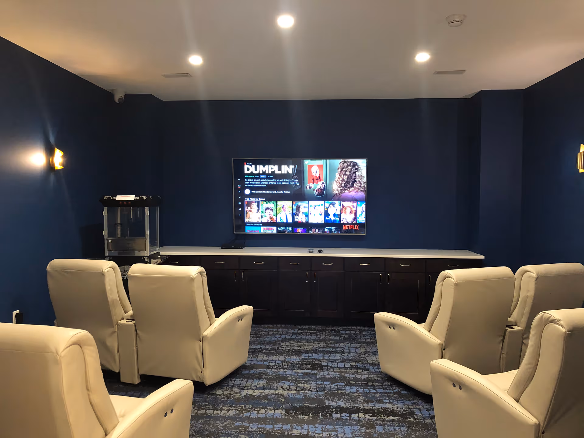A small movie theater room with six beige recliner chairs arranged in two rows facing a wall-mounted flat-screen TV displaying the movie 'Dumplin''. The walls are painted dark blue, and there are two wall sconces providing soft lighting. A popcorn machine is placed on a counter beneath the TV.