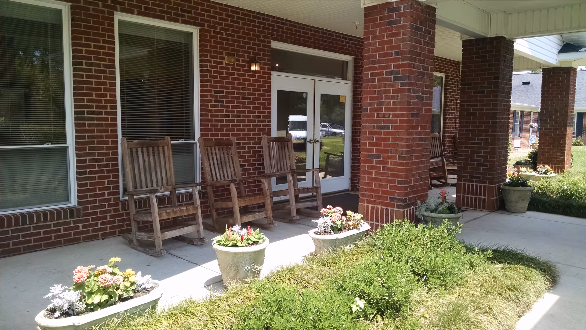 Covered outdoor patio area with four wooden rocking chairs lined up against a brick wall with windows and a glass door. Several flower pots with colorful flowers are placed along the edge of the patio, and green bushes and grass are visible in the foreground.