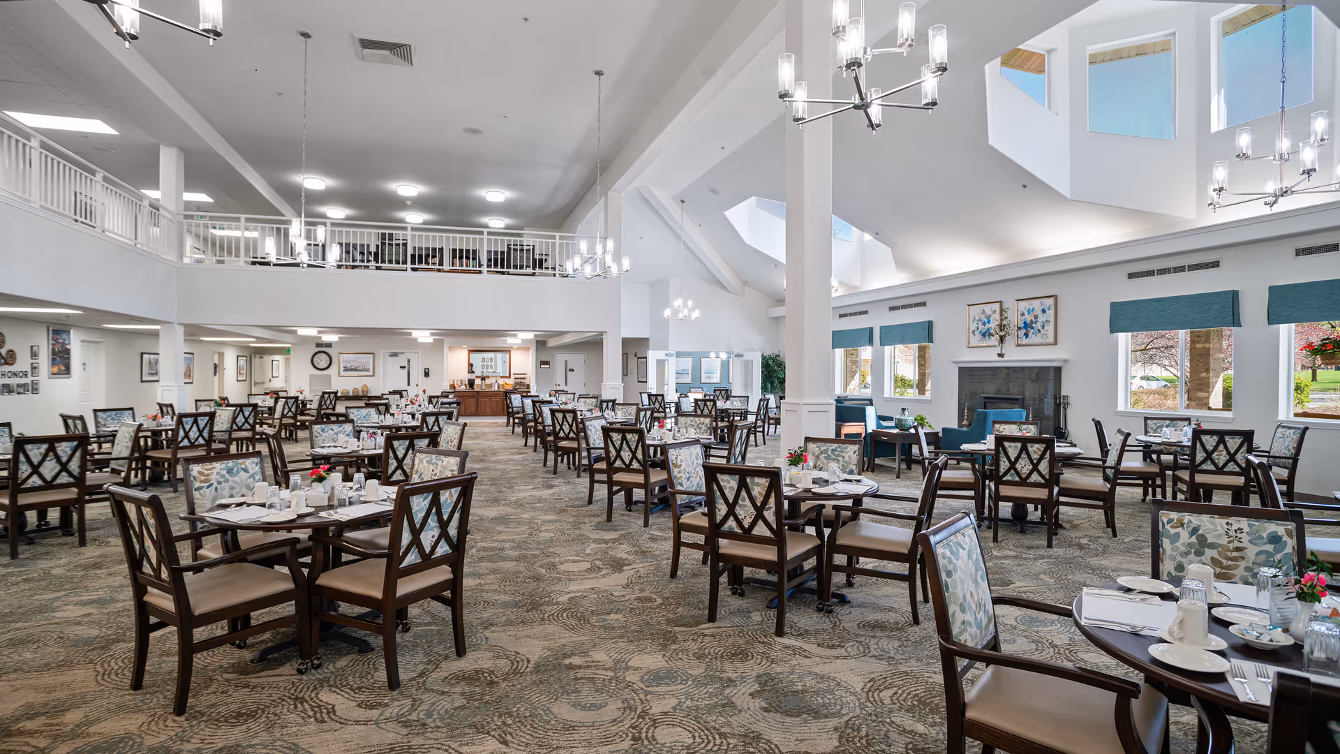 Spacious dining room in a retirement living facility with multiple round tables and chairs arranged neatly. The room features high ceilings with modern chandeliers, large windows allowing natural light, and a cozy seating area with armchairs near a fireplace. The decor includes floral artwork and patterned carpet.