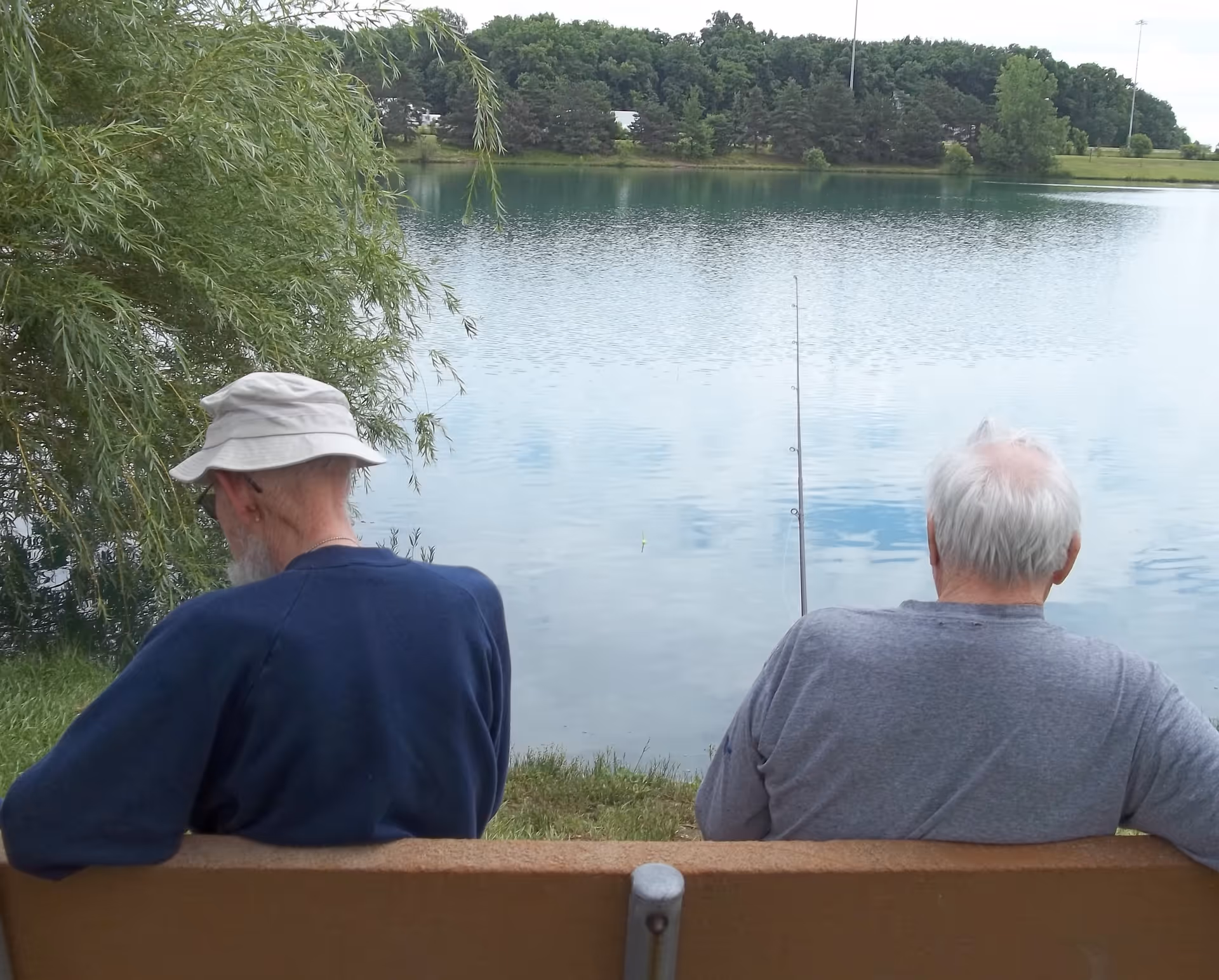 Two elderly men sitting on a bench by a lake, one wearing a white hat and blue shirt, the other in a gray shirt, with a fishing rod set up in front of them and trees in the background.