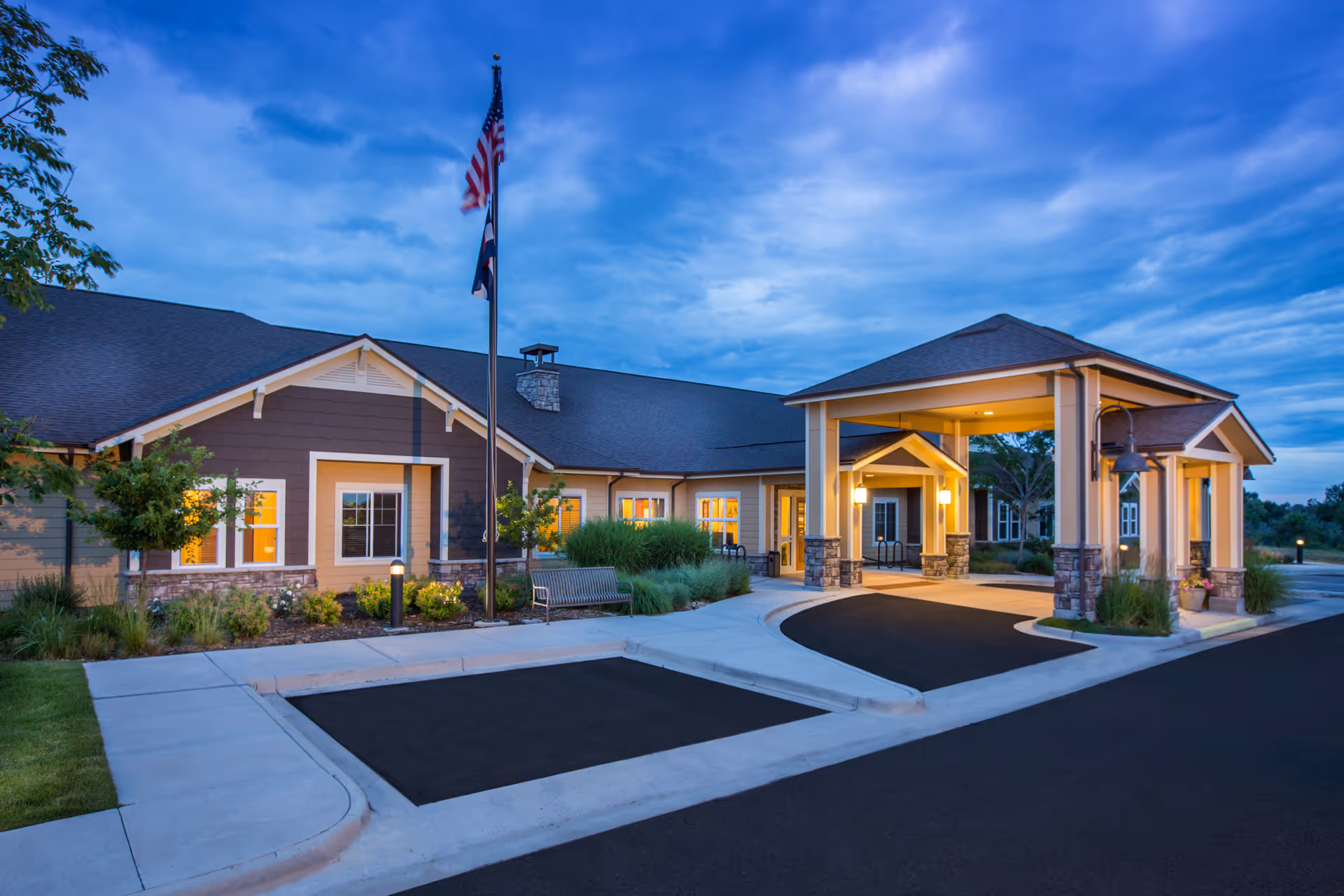 Front exterior of a memory care facility at dusk showing a covered entrance with a flagpole, benches, and landscaping.