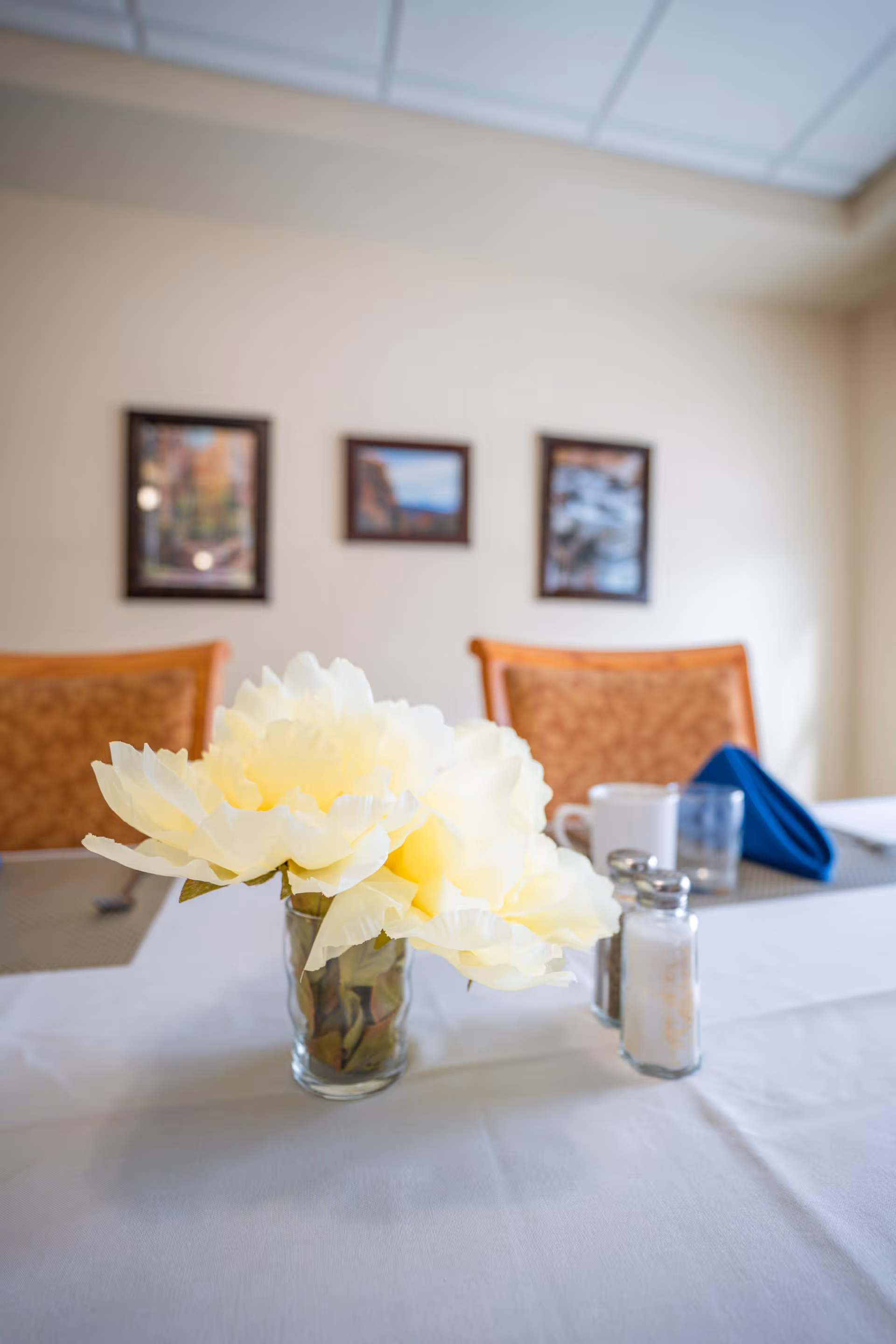 A dining table set with a white tablecloth, a glass vase holding large pale yellow flowers, salt and pepper shakers, a white coffee mug, a glass, and a folded blue napkin. In the background, there are two orange cushioned chairs and three framed pictures on a beige wall.