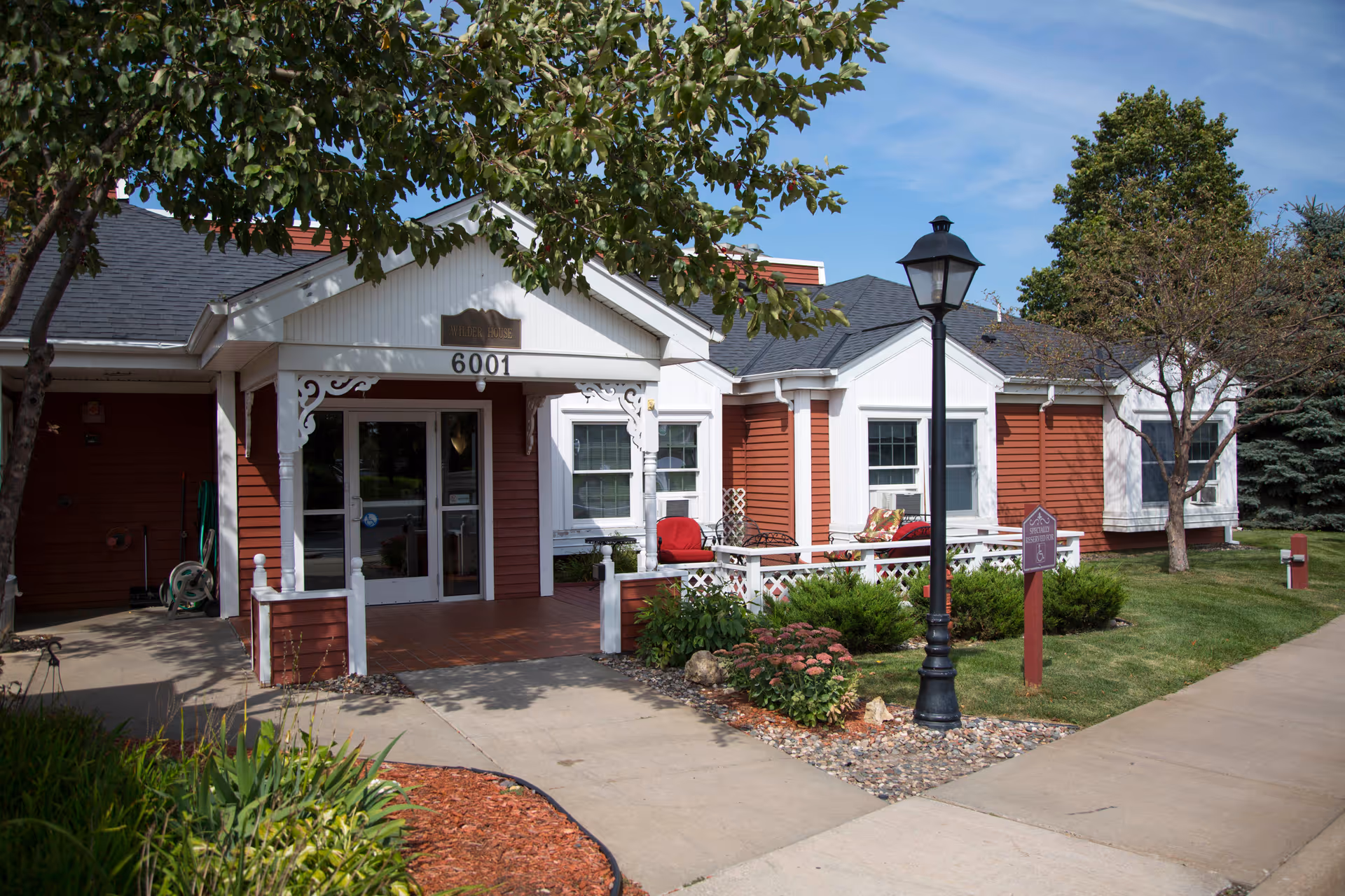 Front exterior of a red-and-white senior living building with a covered entrance numbered 6001, a porch, and a lamppost.