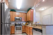Modern kitchen with wooden cabinets, stainless steel refrigerator, microwave, oven, and dishwasher. The countertop is granite with various kitchen items placed on it. The kitchen is well-lit with ceiling lights and has a clean, organized appearance.
