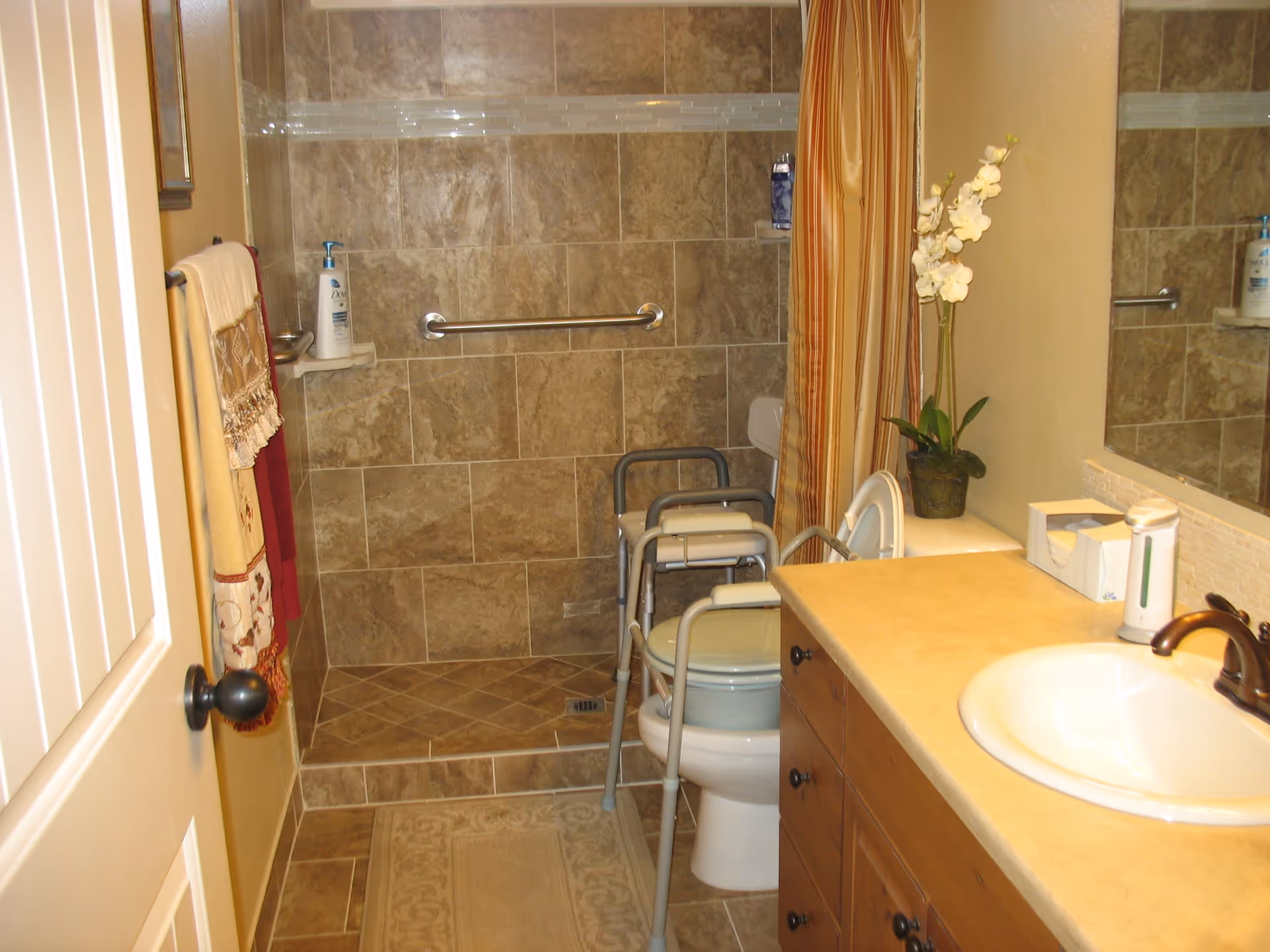 A bathroom with beige tiled walls and floor, featuring a walk-in shower with a grab bar and a shower chair. There is a toilet with a support frame around it, a countertop with a sink, a faucet, a tissue box, a soap dispenser, and a potted plant with white flowers. Towels hang on a rack near the door.