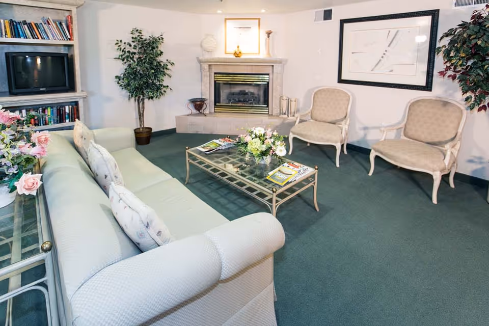 A cozy living room with a light green sofa, two beige armchairs, a glass coffee table with magazines and a flower arrangement, a fireplace with decorative items on the mantel, a built-in bookshelf with books and a TV, and potted plants in the corners.