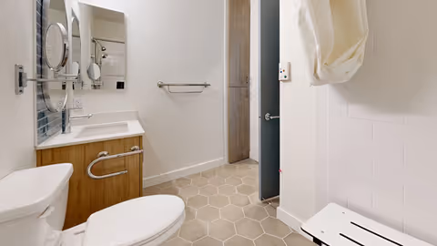 A clean and modern bathroom with a white toilet, a wooden vanity with a white sink, a mirror above the sink, a towel rack on the wall, and a shower area with a white foldable shower seat and a white shower curtain.