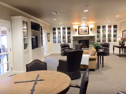 A cozy living room area in an assisted living facility featuring a round wooden table with chairs in the foreground, several armchairs and a sofa arranged around a stone fireplace with a painting above it. Built-in white cabinets with glass doors flank the fireplace, and there are plants and decorative items placed around the room. The space is well-lit with ceiling lights and wall sconces.