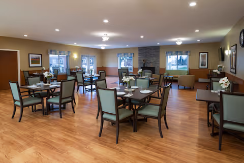 A spacious dining room with multiple wooden tables and chairs arranged neatly. Each table is set with white plates and napkins, and decorated with small flower arrangements. The room has wooden flooring, beige walls with a darker wainscoting, and several windows with blue and white curtains. There is a stone fireplace in the background and a few framed pictures on the walls.