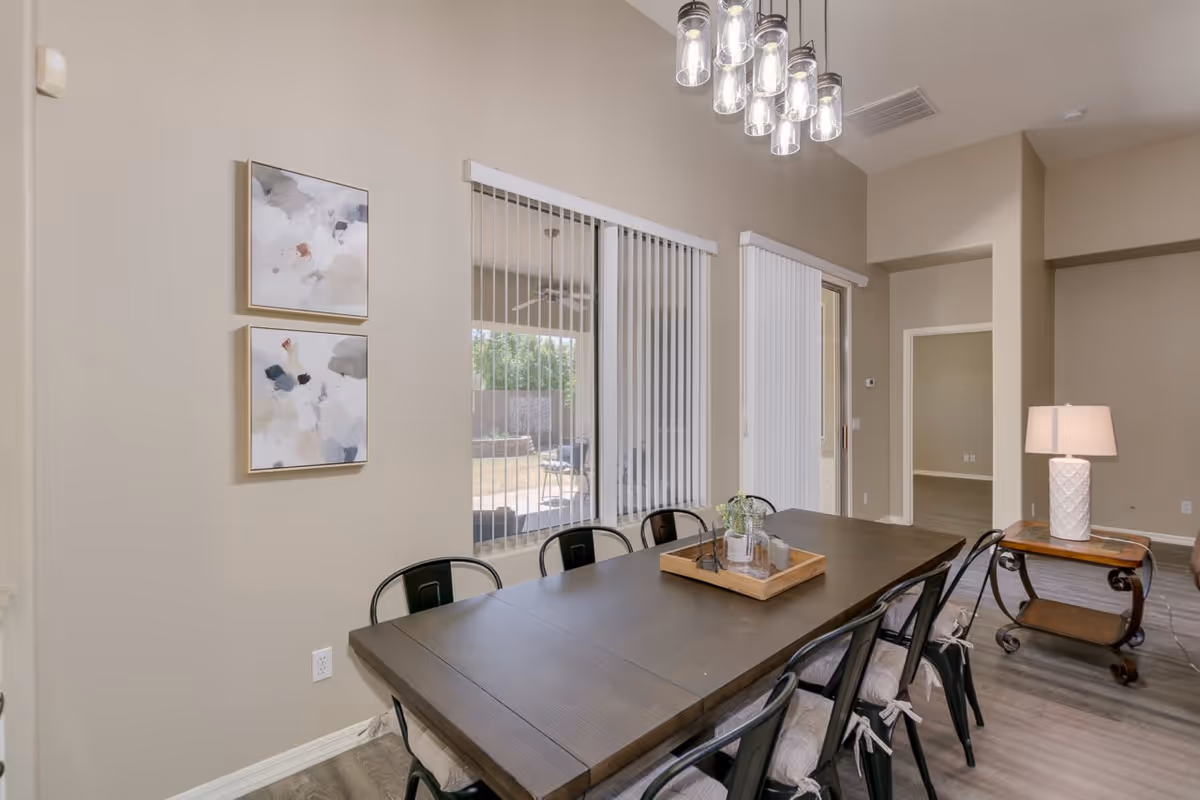 Modern dining room with a long dark wooden table surrounded by black metal chairs, pendant lights, vertical blinds, and wall art.