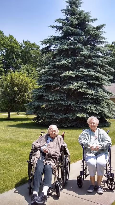 Two elderly women sitting in wheelchairs on a paved path outside on a sunny day. They are in front of a large evergreen tree with green grass and other trees in the background.