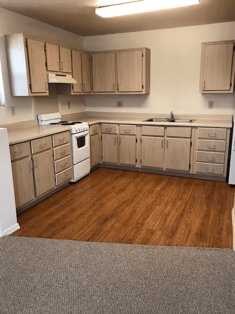 A kitchen with light wood cabinets, a white stove with an oven, a double sink, and beige countertops. The floor is a wood laminate, and the walls are painted white. There is a fluorescent ceiling light and a small window on the left side.