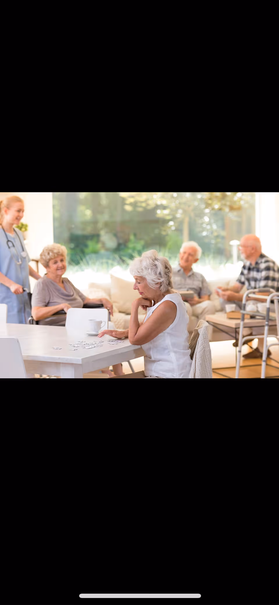 A group of elderly people sitting and interacting in a bright, comfortable common area with large windows. One elderly woman is sitting at a table playing a game, while two elderly men and another woman are seated on a couch in the background. A caregiver in scrubs stands nearby, smiling.