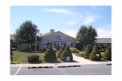 Front entrance of a single-story brick senior living facility with manicured shrubs, an American flag, and parking spaces.