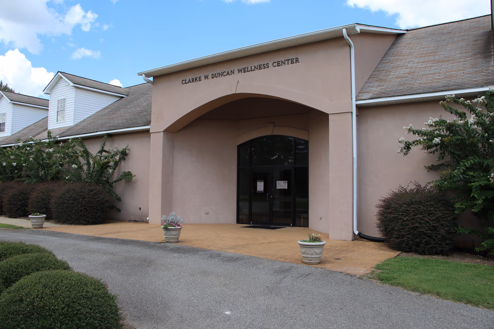 Exterior view of the Clarke W. Duncan Wellness Center building with a beige facade, an arched entrance with glass doors, surrounded by bushes and potted plants, under a partly cloudy sky.