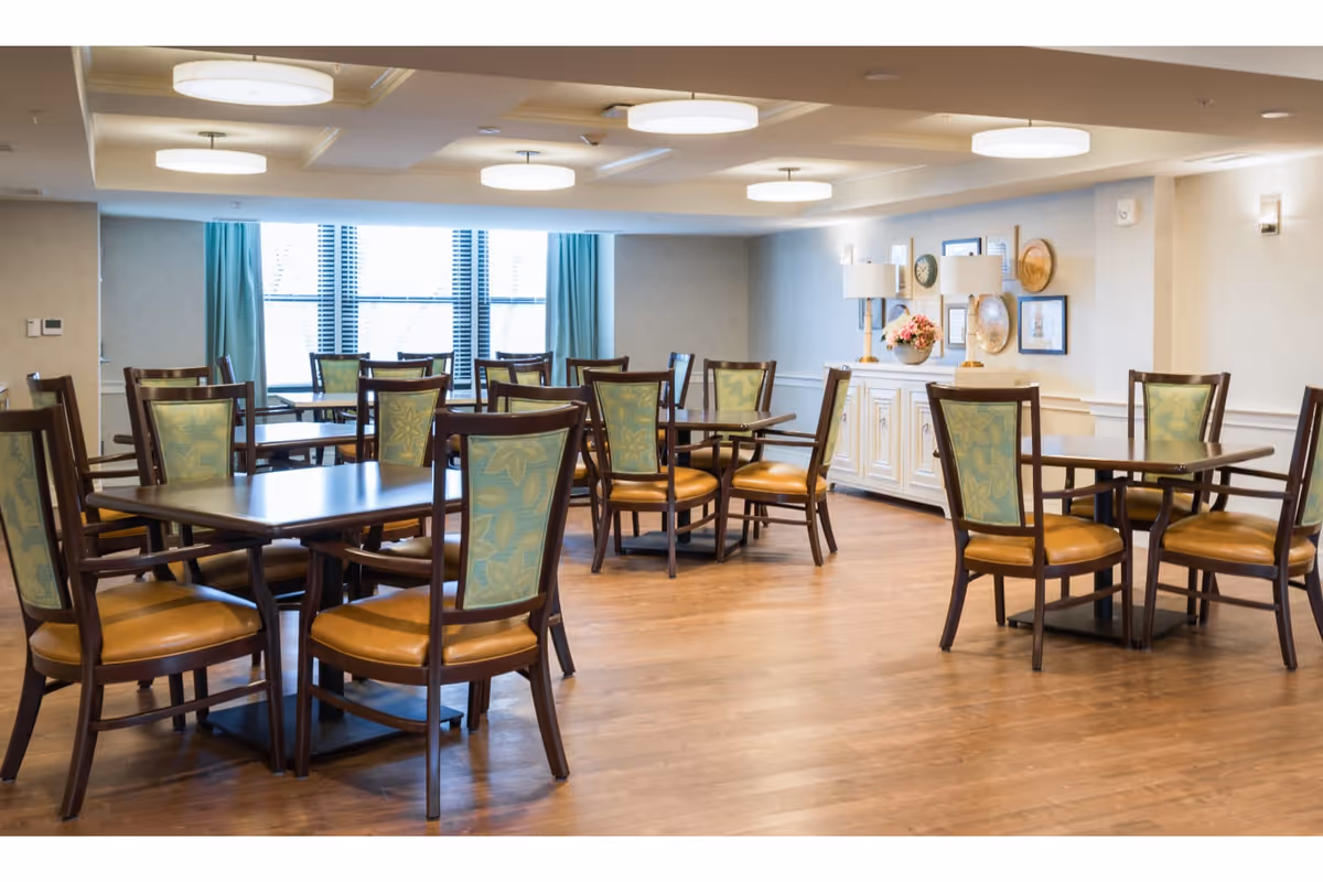 A bright dining room with multiple wooden tables and chairs featuring green patterned backs and tan seats. The room has large windows with blue curtains, wooden flooring, ceiling lights, and a white sideboard decorated with lamps, flowers, and framed pictures on the wall.